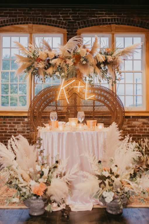 Photo of a sweetheart table with our double throne peacock chair rental, florals, and a lit sign with the couple's initials