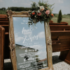 Large mirror with ornate gold frame with white writing at a wedding. Mirror is propped up against outdoor pews.