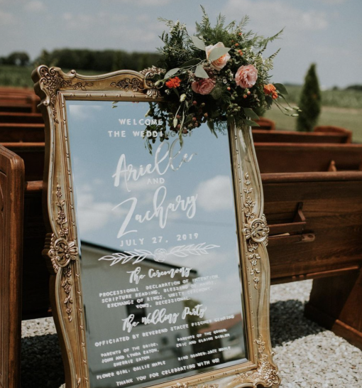 Large mirror with ornate gold frame with white writing at a wedding. Mirror is propped up against outdoor pews.