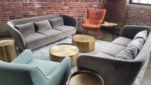 Lounge seating area at a corporate event with two modern gray sofas, an orange wingback arm chair, a seafoam green armchair and gold circular coffee tables