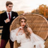 Woman sitting in peacock chair with a man looking at her. Outside in a field in the fall with black fence in the background.