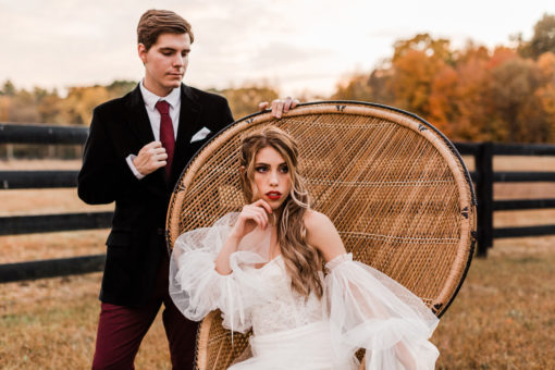 Woman sitting in peacock chair with a man looking at her. Outside in a field in the fall with black fence in the background.