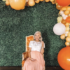 Pregnant woman at her baby shower sitting in an ornate orange armchair against a green backdrop with orange balloons along the edges.