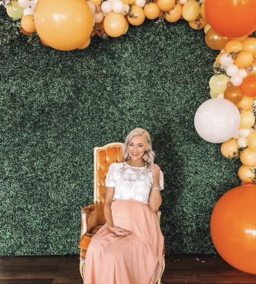 Pregnant woman at her baby shower sitting in an ornate orange armchair against a green backdrop with orange balloons along the edges.