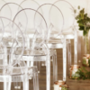 Clear acrylic ghost chairs set up in rows at a wedding ceremony. Florals line the aisle.
