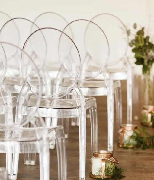 Clear acrylic ghost chairs set up in rows at a wedding ceremony. Florals line the aisle.