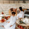 Wives in an outdoor photoshoot. Round coffee table in the foreground and gray sofa in the background.