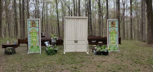 Entrance doors at an outdoor wedding ceremony. Doors are freestanding and closed. Two stain glass panels are to the side with church pews behind.
