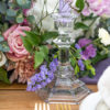 Glass candlestick in a tablescape with purple and pink hued flowers in the background