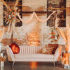 Soft pink velvet sofa with orange pillows with a boho look and florals on gold side tables. Backlit with a brick wall.
