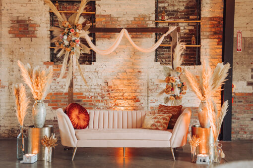 Soft pink velvet sofa with orange pillows with a boho look and florals on gold side tables. Backlit with a brick wall.