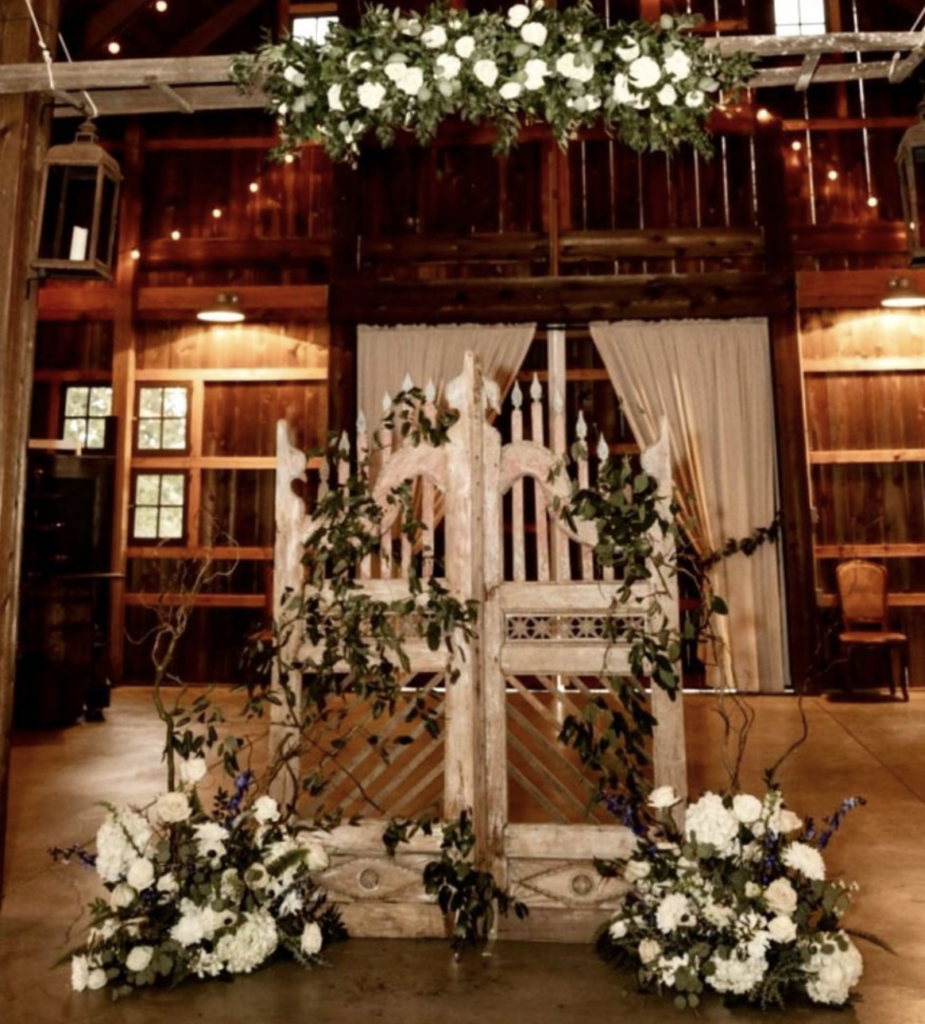 Elaborate carved gate doors used as a backdrop in a wedding ceremony. Florals drape the doors and the ground. Wedding held in a barn.
