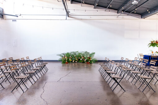 Folding wood and metal bistro chairs arranged in rows facing a middle aisle. Florals at the end again a wall