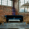 Lounge seating area with blue velvet modern sofa, two rattan barrel chairs, white coffee table, rug, and a large decorative umbrella against a brick wall and industrial windows