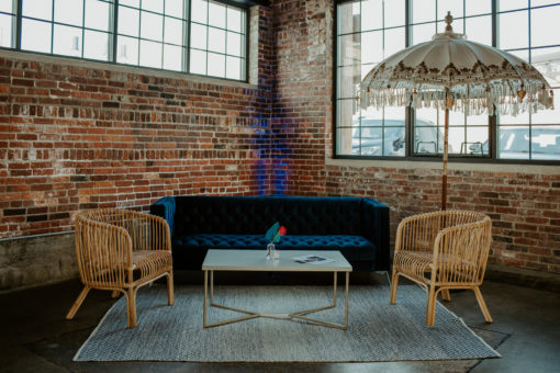 Lounge seating area with blue velvet modern sofa, two rattan barrel chairs, white coffee table, rug, and a large decorative umbrella against a brick wall and industrial windows