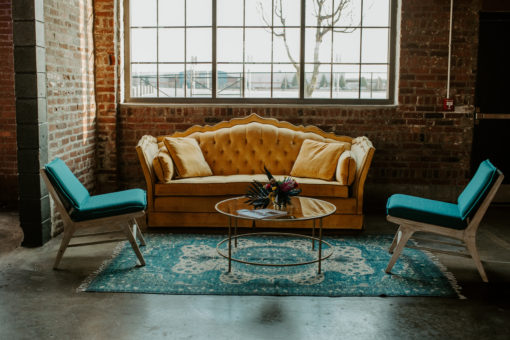 Yellow sofa, two blue chairs, a teal rug, glass round coffee table lit by a giant window.