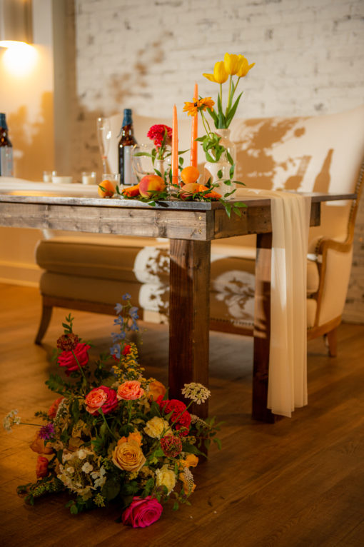 Side view of our wooden farm table used as a sweetheart table in a wedding reception. Florals at the feet and on the top. Loveseat in the back.