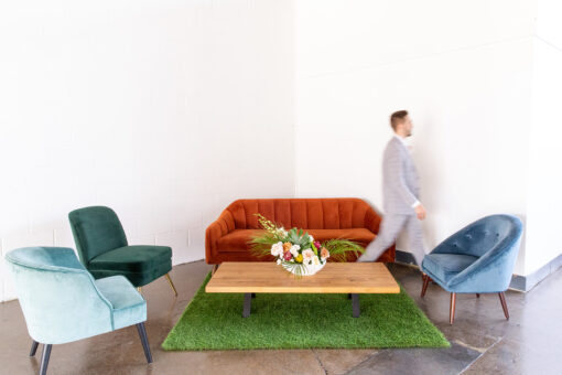 Burnt orange mid century mod sofa with side arm chairs in blue and green. Faux grass carpet under a wooden coffee table.