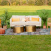 White sofa sitting outside in a field for a photo shoot. Two gold coffee tables sitting on a rug. Two rattan chairs on the right side and a brown pouf on the left.