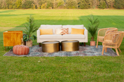 White sofa sitting outside in a field for a photo shoot. Two gold coffee tables sitting on a rug. Two rattan chairs on the right side and a brown pouf on the left.