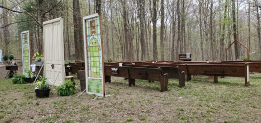 Freestanding doors and stained glass create the entrance to this outdoor wedding ceremony with church pews. Surrounded by woods