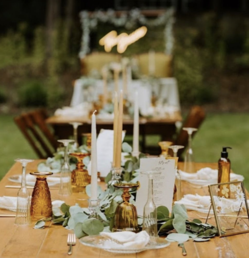 Outdoor dinner party. Wooden long farm table with candles, greenery, and amber mismatched goblets.