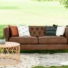 Brown leather sofa sitting outside in a field for a styled shoot. White, black, and orange pillows on the couch. Rattan coffee table. Two rugs, and a plant set up in a lounge seating setting.