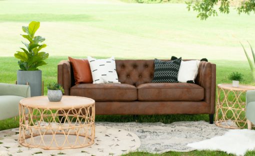 Brown leather sofa sitting outside in a field for a styled shoot. White, black, and orange pillows on the couch. Rattan coffee table. Two rugs, and a plant set up in a lounge seating setting.