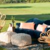 Angled view of a lounge setting area outside on grass. Navy curved modern sofa with pink pillows. Two macramé poufs in front. Rattan chairs on each side. Sitting on a rug.