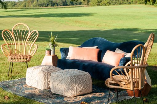 Angled view of a lounge setting area outside on grass. Navy curved modern sofa with pink pillows. Two macramé poufs in front. Rattan chairs on each side. Sitting on a rug.