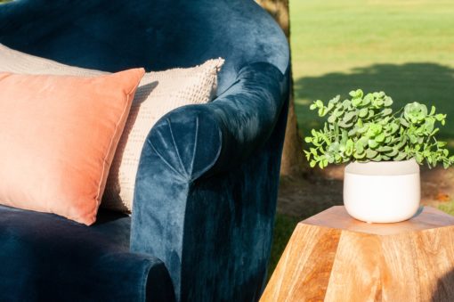 Close up of navy sofa with curved arm. Pink pillows. Wooden geometric side table with small plant in a white vase in a styled shoot outdoor lounge setting.