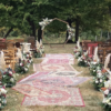 Outdoor wedding ceremony with mismatched rugs in the aisle, geometric wooden arch with florals and mismatched wooden chairs for guest seating.
