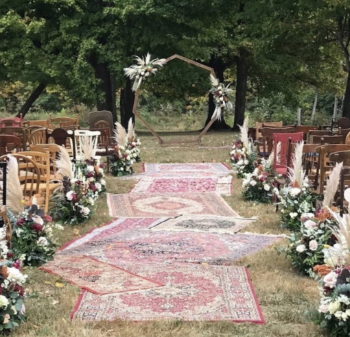 Outdoor wedding ceremony with mismatched rugs in the aisle, geometric wooden arch with florals and mismatched wooden chairs for guest seating.