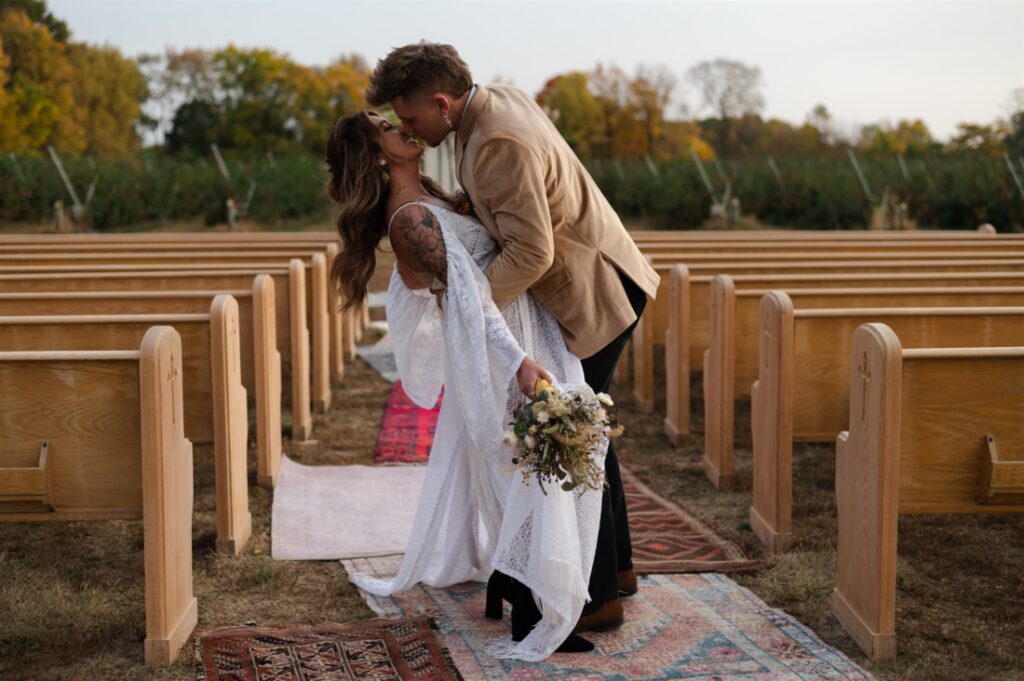 Couple kissing in an outdoor wedding with church pews and rugs for the aisle