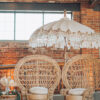 Two light rattan peacock chairs with white cushions under a boho white and gold umbrella. Brick background with a natural light window.