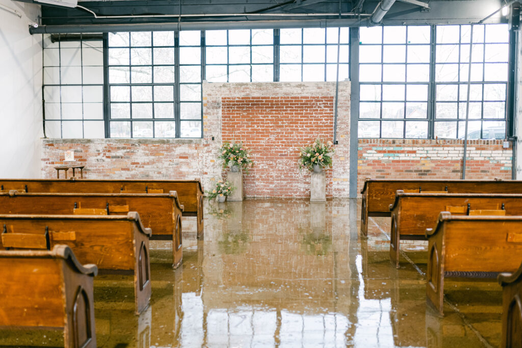 Industrial venue set up for wedding. Bank of windows up front with exposed brick wall. Pews set up for ceremony with aisle down the center.