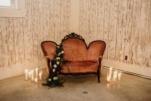 Pink victorian velvet loveseat draped with florals and surrounded by candles. White barn walls in the background