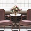 Vintage purple velvet armchairs with wooden arms. In between the chairs is a dark wood vintage side table with flowers in a vase. Naturally backlit by a large window.