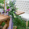 Ivory long settee with no arms used at a wedding reception photo shoot head table. Farm table with purple florals and greenery. Outdoors on grass