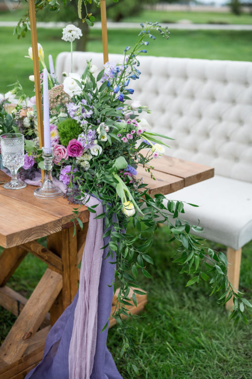 Ivory long settee with no arms used at a wedding reception photo shoot head table. Farm table with purple florals and greenery. Outdoors on grass