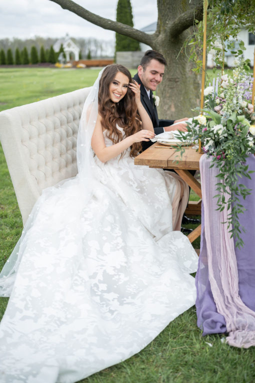 A young couple sitting on the settee at a wedding sweetheart table. On the grass, tree in background.