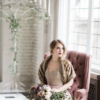 Styled photo shoot with woman sitting on a muted purple armchair. Large window with natural light behind. Floor candelabra in background.