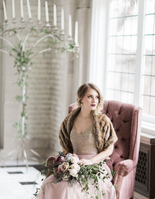 Styled photo shoot with woman sitting on a muted purple armchair. Large window with natural light behind. Floor candelabra in background.