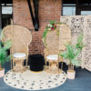 Peacock chairs on a round jute rug. Plants and a black round ottoman. Wooden backdrop behind the chairs.