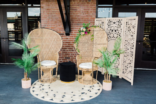 Peacock chairs on a round jute rug. Plants and a black round ottoman. Wooden backdrop behind the chairs.