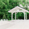 White wire chairs set up for an outdoor wedding ceremony. Green trees in background with a small covered structure