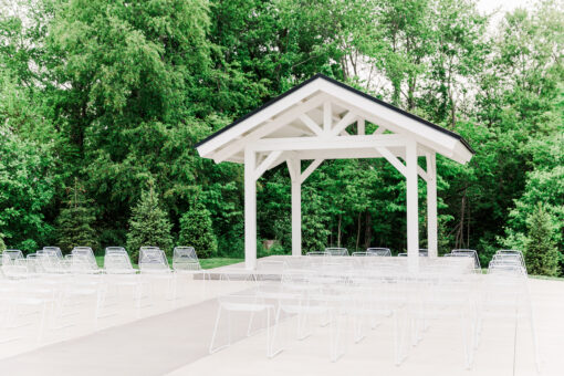 White wire chairs set up for an outdoor wedding ceremony. Green trees in background with a small covered structure