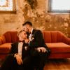 One man kissing the forehead of another man while sitting on a burnt orange mid-century modern sofa.