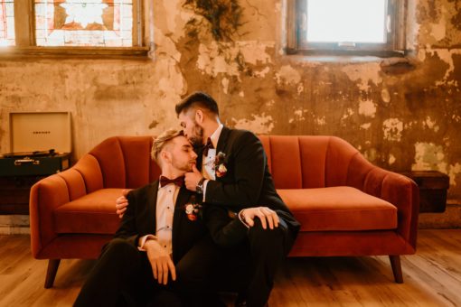 One man kissing the forehead of another man while sitting on a burnt orange mid-century modern sofa.