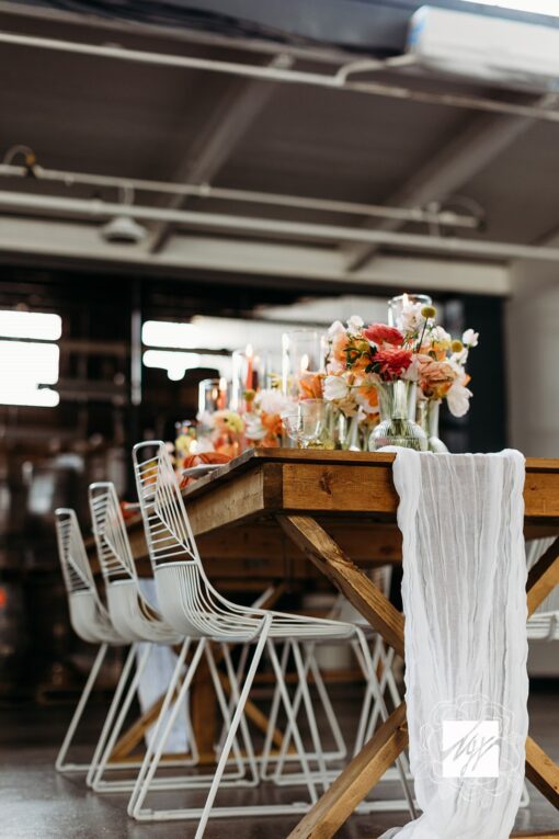 Wedding reception setup with farmtable and white wire chairs. White drape on the table with florals.